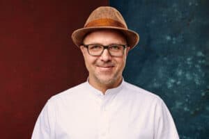 Mann mit Brille und Hut, lächelnd, vor buntem Hintergrund.
